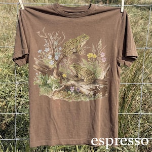 May include: Espresso-colored t-shirt featuring a detailed illustration of two frogs on a log surrounded by wildflowers. The frogs are green and brown, and the shirt has a classic crew neck design. The word "espresso" is printed at the bottom.