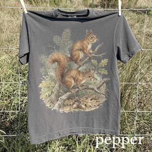 May include: A gray t-shirt featuring a detailed illustration of two squirrels perched on a tree branch with leaves and acorns. The word "pepper" is printed at the bottom of the shirt.