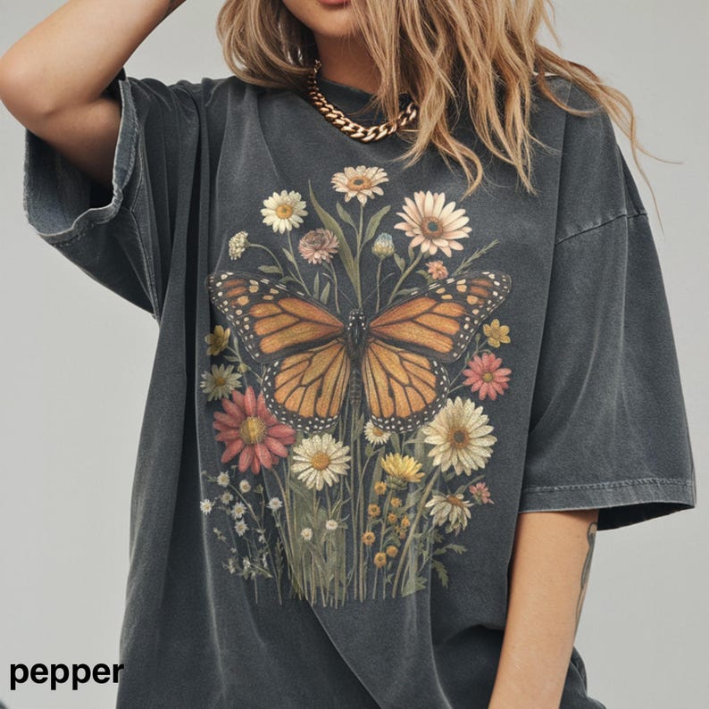 Puede incluir: Camiseta gris oscuro de gran tama&ntilde;o con una gran mariposa con alas naranjas y negras, rodeada de flores coloridas. La camiseta tiene un aspecto vintage y descolorido. La palabra "pepper" est&aacute; impresa en la parte inferior izquierda.