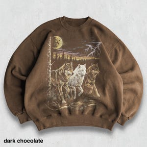 May include: Dark chocolate-colored sweatshirt featuring a graphic of three wolves running under a moonlit sky with lightning. The text "dark chocolate" is printed at the bottom. The sweatshirt has a crew neck and long sleeves.