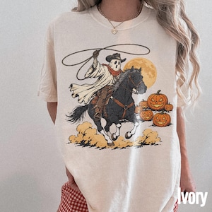 Könnte beinhalten: Ein elfenbeinfarbenes Kurzarm-T-Shirt mit einem Grafikdruck eines Geistes, der einen Cowboyhut und ein Halstuch trägt, auf einem schwarzen Pferd reitet und ein Lasso schwingt, vor einem großen gelben Mond und orangefarbenen Kürbissen.
