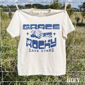 May include: Ivory-colored t-shirt with the words "GRACE ROCKY SAVE STARS" in blue block letters. The shirt is hanging on a clothesline in front of a grassy field. The shirt has short sleeves and a crew neck.