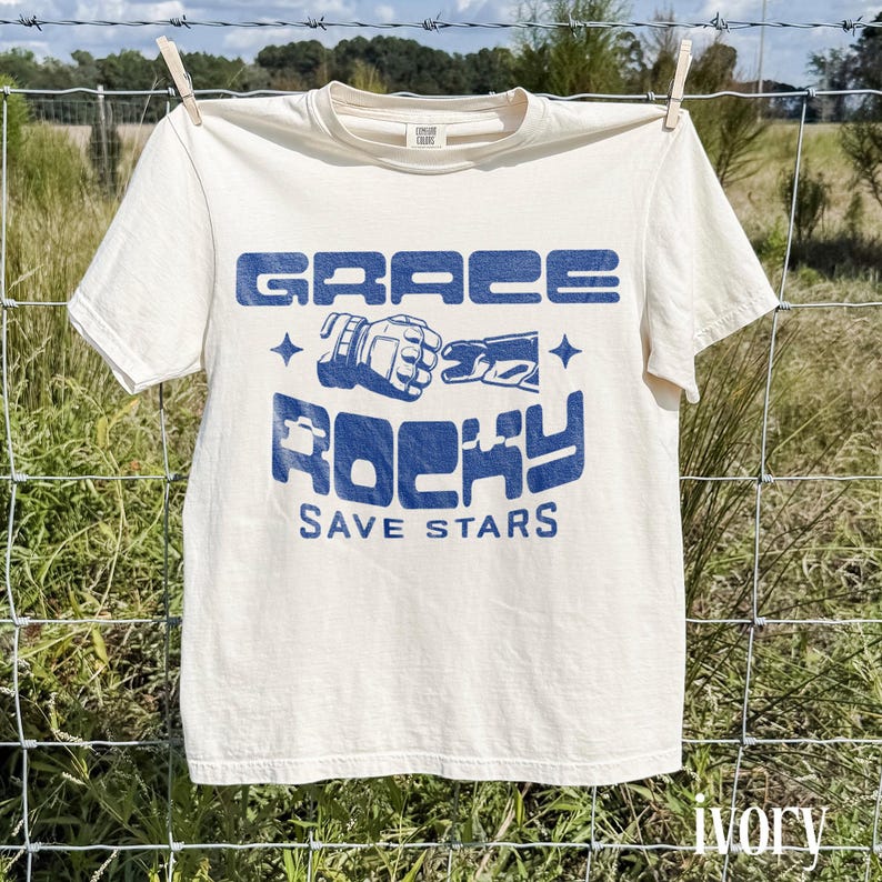 May include: Ivory-colored t-shirt with the words "GRACE ROCKY SAVE STARS" in blue block letters. The shirt is hanging on a clothesline in front of a grassy field. The shirt has short sleeves and a crew neck.