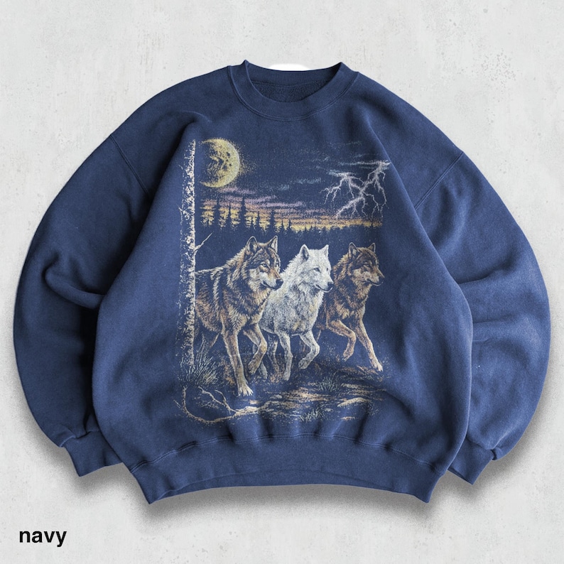 May include: Navy blue sweatshirt featuring a graphic of three wolves running through a forest under a moonlit sky with lightning. The text "navy" is printed at the bottom. The sweatshirt has a crew neck and long sleeves.