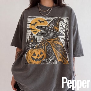 May include: A charcoal gray short-sleeve t-shirt featuring a graphic print of a crow wearing a witch hat and cape, standing next to a jack-o'-lantern. The background of the print shows a full moon, flying bats, and a silhouette of a castle.