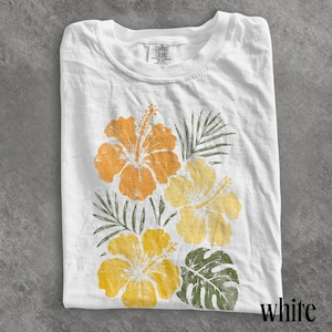 May include: A white short-sleeve t-shirt featuring a graphic print of three hibiscus flowers in shades of orange and yellow, accompanied by green palm and monstera leaves. The inner neck label displays the text Comfort Colors.