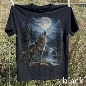May include: Black t-shirt featuring a detailed graphic of a wolf howling at the moon in a forest setting. The design includes a waterfall and mountains under a bright, full moon. The word "black" is printed at the bottom right.