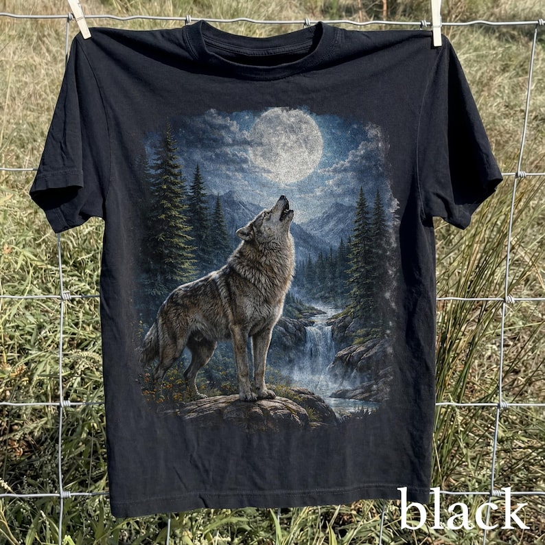 May include: Black t-shirt featuring a detailed graphic of a wolf howling at the moon in a forest setting. The design includes a waterfall and mountains under a bright, full moon. The word "black" is printed at the bottom right.