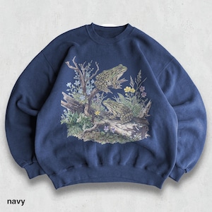 May include: Navy blue sweatshirt featuring a detailed graphic of two frogs perched on a log with foliage and flowers. The design includes a tree branch and various plants. The word "navy" is printed at the bottom.