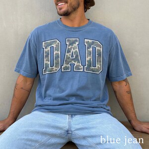 以下が含まれることがあります： 迷彩柄の大きなブロック文字で「DAD」と書かれた青いTシャツ。明るい青色のジーンズと合わせています。画像の下部には「blue jean」という文字があります。