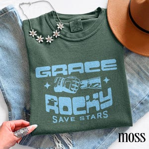 May include: A moss green t-shirt with the text "GRACE ROCKY SAVE STARS" in light blue. The shirt features a graphic of two fists bumping. A silver necklace with flower-shaped pendants and a brown hat are also visible.
