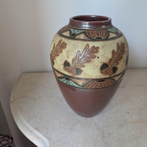 May include: A ceramic vase with a rounded body and a narrow neck. The lower portion is solid brown, while the upper section features a cream-colored band decorated with embossed brown oak leaves and acorns, accented by green geometric patterns.