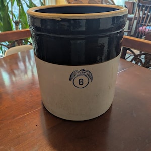 May include: A cylindrical stoneware crock featuring a two-tone design with a dark brown glazed upper section and a light beige lower body. A blue stamped graphic of an eagle perched above the number 6 is centered on the front of the vessel.