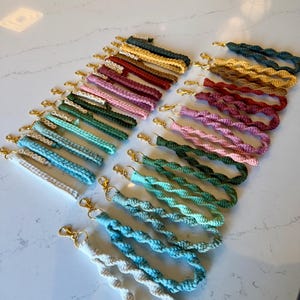 May include: A collection of macrame wrist keychains in various colors, including blue, green, pink, and brown. Each keychain features a gold-colored clasp and a woven design. Some keychains have names spelled out in small white beads.