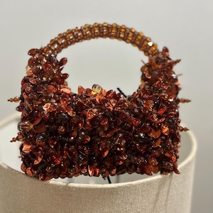 May include: A small, beaded handbag in shades of amber and brown. The bag is covered in small, irregular beads, with a rounded handle made of smaller, round beads. The bag is sitting on a neutral-colored surface.