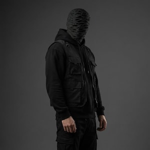 Op de afbeelding: Een persoon draagt een zwarte hoodie, een zwart vest met meerdere zakken en een zwarte cargobroek. De persoon draagt ook een zwart masker met een uniek, gestructureerd patroon. De achtergrond is effen grijs.