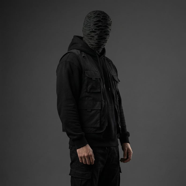 Op de afbeelding: Een persoon draagt een zwarte hoodie, een zwart vest met meerdere zakken en een zwarte cargobroek. De persoon draagt ook een zwart masker met een uniek, gestructureerd patroon. De achtergrond is effen grijs.