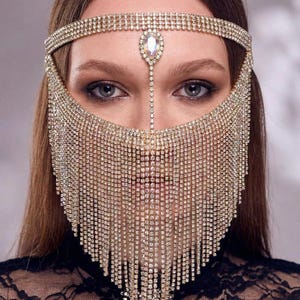 May include: A glamorous headpiece featuring a sparkling rhinestone headband and a face veil. The veil cascades down in delicate strands, creating a dazzling effect. A large, teardrop-shaped rhinestone adorns the forehead. The headpiece is perfect for special occasions.