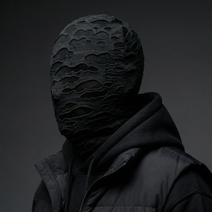 Op de afbeelding: Een persoon draagt een zwarte hoodie, een zwart vest en een zwart gezichtsmasker. Het gezichtsmasker heeft een versleten, gescheurd ontwerp, dat het gezicht eronder onthult. De algehele esthetiek is donker en modern.