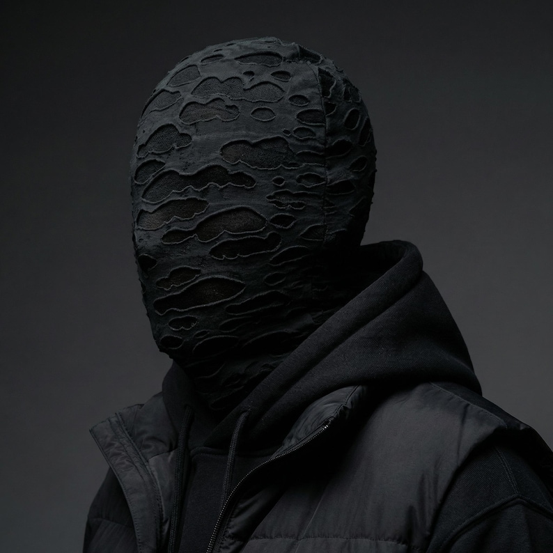 Op de afbeelding: Een persoon draagt een zwarte hoodie, een zwart vest en een zwart gezichtsmasker. Het gezichtsmasker heeft een versleten, gescheurd ontwerp, dat het gezicht eronder onthult. De algehele esthetiek is donker en modern.