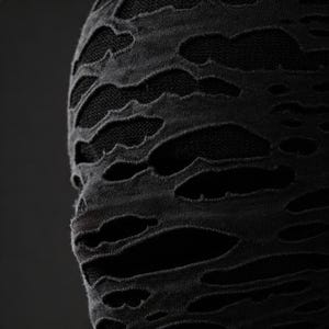 Op de afbeelding: Close-up van een zwarte stof met een distressed, gescheurd ontwerp. De stof heeft meerdere onregelmatige openingen, die een donkerdere, onderliggende laag onthullen. De textuur lijkt zacht en licht versleten, wat een uniek visueel effect cre&euml;ert.