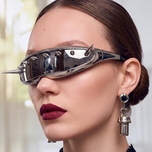 Könnte beinhalten: Silberfarbene futuristische Brille mit mehreren Stacheln und verspiegelten Gläsern. Die Brille hat ein schlankes, umlaufendes Design. Das Model trägt dunkelroten Lippenstift und passende Ohrringe.