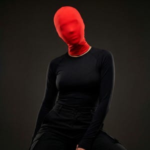 Op de afbeelding: Een persoon draagt een rode volledige gezichtsbedekking en een zwart shirt met lange mouwen en een zwarte broek. De rode bedekking verbergt het gezicht, waardoor een mysterieuze en anonieme uitstraling ontstaat. De outfit staat tegen een donkergrijze achtergrond.