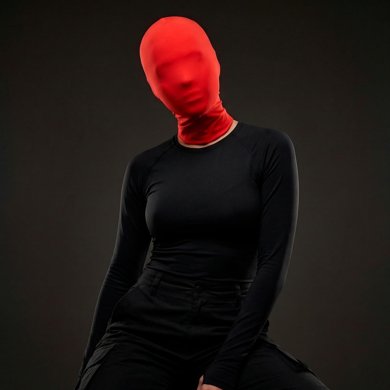 Op de afbeelding: Een persoon draagt een rode volledige gezichtsbedekking en een zwart shirt met lange mouwen en een zwarte broek. De rode bedekking verbergt het gezicht, waardoor een mysterieuze en anonieme uitstraling ontstaat. De outfit staat tegen een donkergrijze achtergrond.