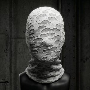 Op de afbeelding: Een wit stoffen masker met een versleten, gescheurd ontwerp, dat het hele hoofd en de nek bedekt. Het masker wordt getoond op een zwarte mannequinhoofd tegen een donkere, industri&euml;le achtergrond.