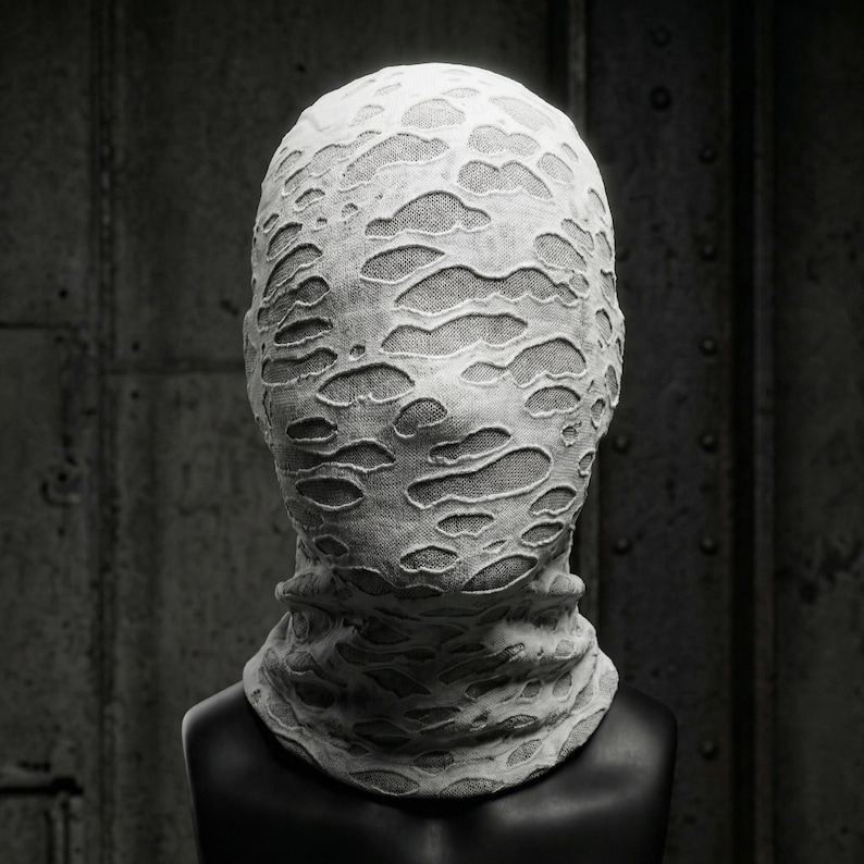 Op de afbeelding: Een wit stoffen masker met een versleten, gescheurd ontwerp, dat het hele hoofd en de nek bedekt. Het masker wordt getoond op een zwarte mannequinhoofd tegen een donkere, industri&euml;le achtergrond.