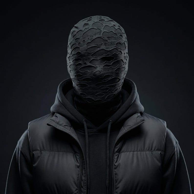Op de afbeelding: Een persoon draagt een zwart masker met een distressed, getextureerd patroon, een zwarte hoodie, een zwart gewatteerd vest en een zwarte coltrui. De afbeelding is tegen een donkere achtergrond geplaatst, waardoor een monochromatische esthetiek ontstaat.