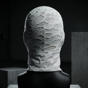 Op de afbeelding: Een witte bivakmuts met een distressed, getextureerd ontwerp. De stof heeft talrijke onregelmatige uitgesneden vormen, waardoor een lichter gekleurd materiaal eronder zichtbaar wordt. De bivakmuts wordt getoond op een zwarte mannequinhoofd tegen een donkere achtergrond.