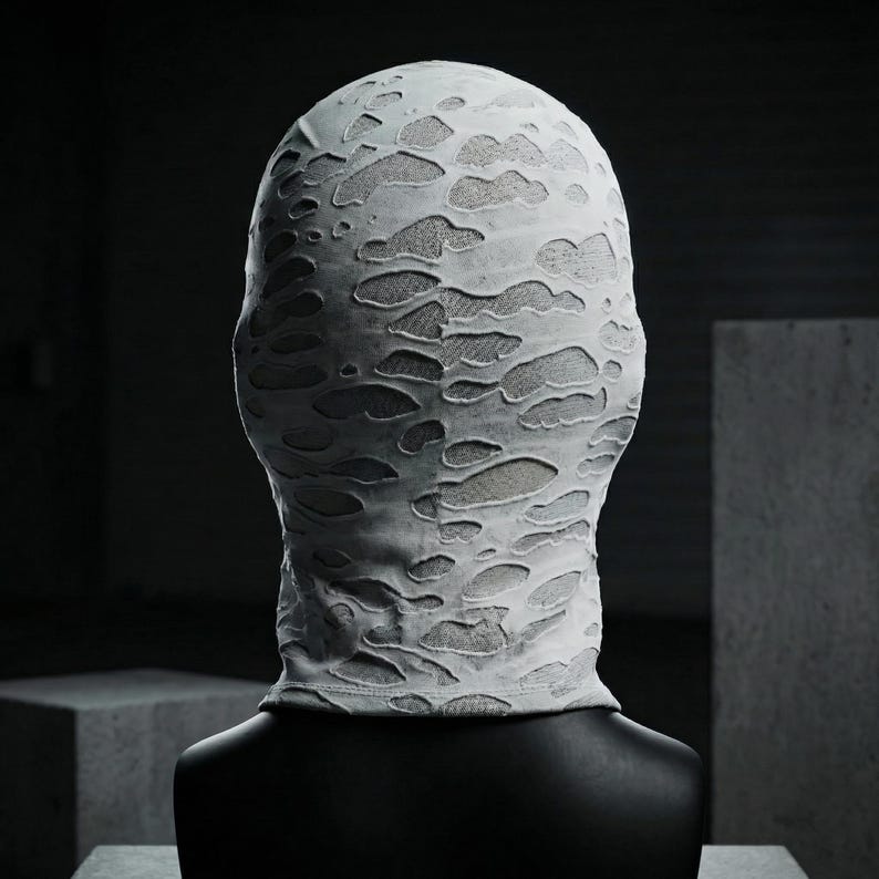 Op de afbeelding: Een witte bivakmuts met een distressed, getextureerd ontwerp. De stof heeft talrijke onregelmatige uitgesneden vormen, waardoor een lichter gekleurd materiaal eronder zichtbaar wordt. De bivakmuts wordt getoond op een zwarte mannequinhoofd tegen een donkere achtergrond.