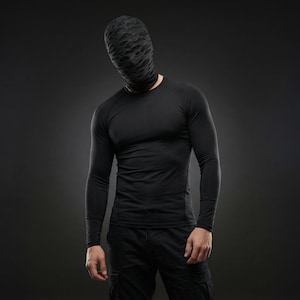 Op de afbeelding: Een persoon draagt een zwart shirt met lange mouwen en een zwarte broek. De persoon draagt ook een zwarte hoofdbedekking met een patroon. De achtergrond is donkergrijs.