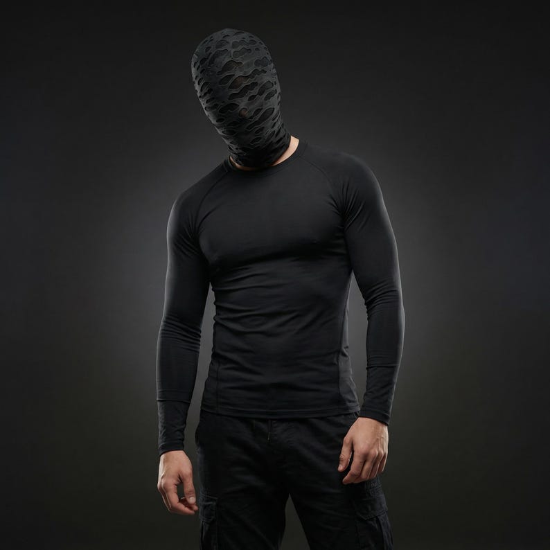 Op de afbeelding: Een persoon draagt een zwart shirt met lange mouwen en een zwarte broek. De persoon draagt ook een zwarte hoofdbedekking met een patroon. De achtergrond is donkergrijs.