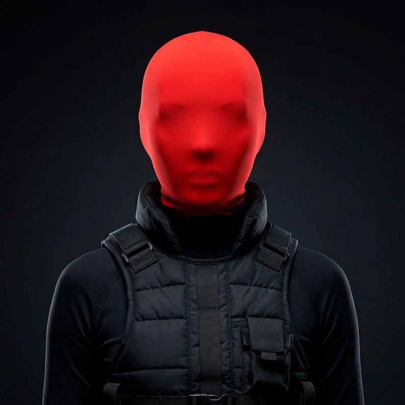 Op de afbeelding: Een persoon draagt een felrode volledige gezichtsbedekking, een zwarte coltrui en een zwart tactisch vest. Het vest heeft meerdere zakken en bandjes. De achtergrond is donkergrijs.