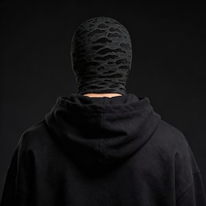 Op de afbeelding: Een zwarte bivakmuts met een vervaagd, wolkenachtig patroon bedekt het hoofd en de nek. De persoon draagt een zwarte hoodie. De achtergrond is effen zwart.