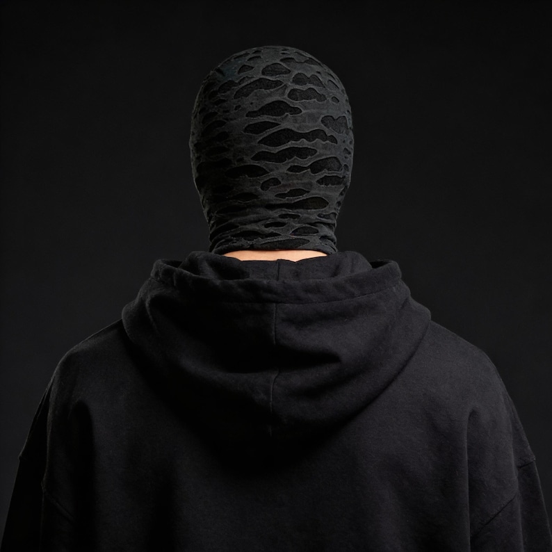 Op de afbeelding: Een zwarte bivakmuts met een vervaagd, wolkenachtig patroon bedekt het hoofd en de nek. De persoon draagt een zwarte hoodie. De achtergrond is effen zwart.