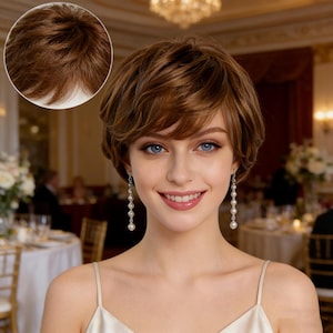 May include: A woman with blue eyes and a warm smile, wearing a short, layered brown wig. The wig has a natural look with varying shades of brown. She is wearing pearl earrings and a cream-colored dress. The background is a blurred interior with a chandelier.