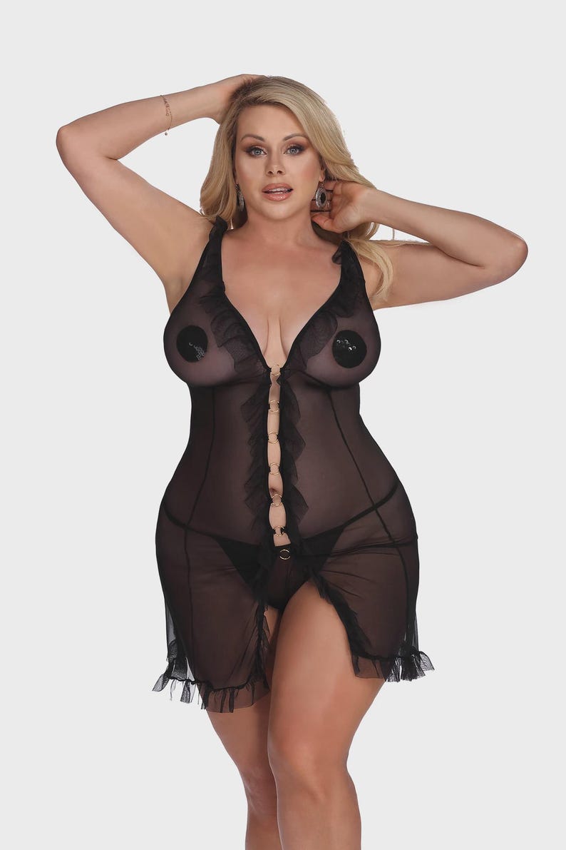 May include: Black lingerie set featuring a sheer babydoll dress with ruffled trim and a front closure. The set includes a matching thong. The dress has a plunging neckline and a high slit. The model is wearing nipple covers.