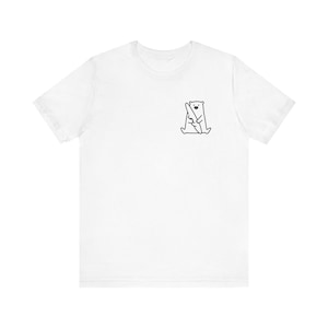 Puede incluir: Camiseta blanca con un pequeño dibujo minimalista en negro de un oso saludando. El gráfico está situado en la parte superior izquierda del pecho. La camiseta tiene mangas cortas y cuello redondo.