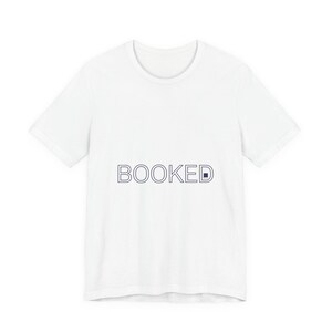 Puede incluir: Camiseta blanca con la palabra "BOOKED" impresa en azul marino. El texto está centrado en la parte delantera. La camiseta tiene mangas cortas y cuello redondo.