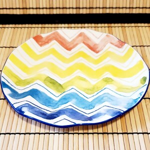 May include: A colorful, oval-shaped ceramic plate with a zig-zag pattern in red, yellow, green, and blue. The plate has a white background and a blue rim. The plate is on a bamboo mat.