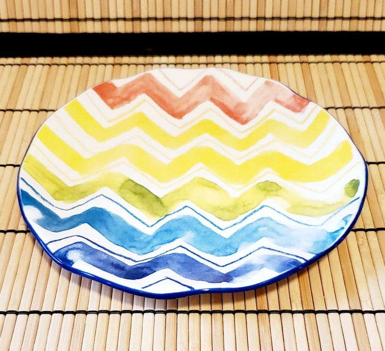 May include: A colorful, oval-shaped ceramic plate with a zig-zag pattern in red, yellow, green, and blue. The plate has a white background and a blue rim. The plate is on a bamboo mat.