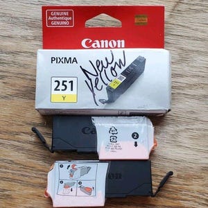 May include: A Canon Pixma 251 Y ink cartridge in its original white and red box with the handwritten text New Yellow on the front. Two additional black ink cartridges are included, each featuring a protective plastic film with printed instructions.