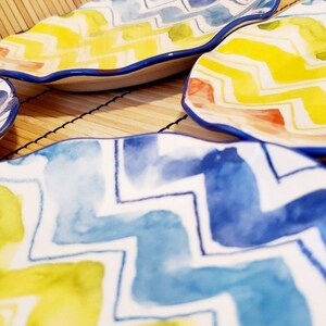 May include: A collection of decorative ceramic plates with a wavy edge. The plates feature a watercolor design with yellow, blue, and green zigzag patterns. The rims are outlined in blue. The plates are arranged on a bamboo mat.