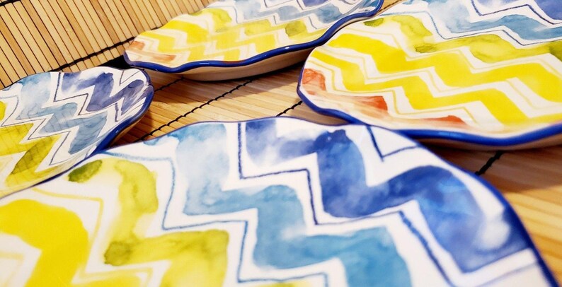 May include: A collection of decorative ceramic plates with a wavy edge. The plates feature a watercolor design with yellow, blue, and green zigzag patterns. The rims are outlined in blue. The plates are arranged on a bamboo mat.