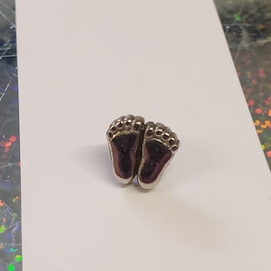May include: A small, metallic silver-toned pin shaped like a pair of baby feet. The design features a smooth, reflective surface with clearly defined toes and a rounded heel shape, presented as a singular decorative accessory item.