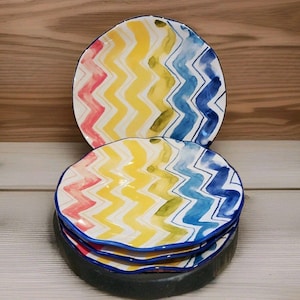 May include: A stack of colorful ceramic plates with a wavy, zig-zag pattern in yellow, blue, pink, and green. The plates have a blue rim and are stacked on a dark gray base. The background is a light wood.