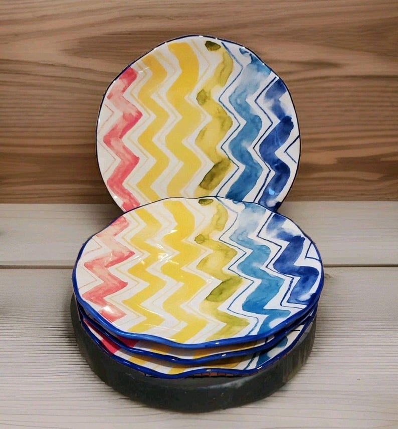 May include: A stack of colorful ceramic plates with a wavy, zig-zag pattern in yellow, blue, pink, and green. The plates have a blue rim and are stacked on a dark gray base. The background is a light wood.